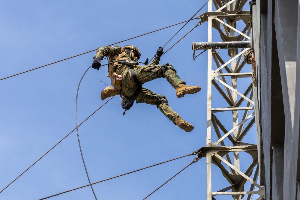 A military personnel in full combat gear and camouflage uniform is performing a rappelling or fast-rope descent from a tall metal tower structure. The soldier is suspended mid-air, connected to ropes, with legs spread and body horizontal against a clear blue sky. A second individual is visible on the tower platform, likely serving as a safety observer or instructor.