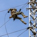 A military personnel in full combat gear and camouflage uniform is performing a rappelling or fast-rope descent from a tall metal tower structure. The soldier is suspended mid-air, connected to ropes, with legs spread and body leaning back in a controlled descent position. A second individual is visible on the tower platform, likely serving as a safety observer or instructor.