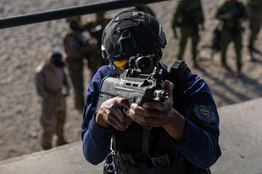 A military or law enforcement operator in the foreground is aiming a compact bullpup-style rifle while wearing tactical gear including a black helmet, yellow-tinted protective eyewear, and a blue uniform with a shoulder patch. Several other personnel in mixed uniforms are visible in the blurred background, suggesting a training exercise or range activity. The setting appears to be an outdoor training facility or range.