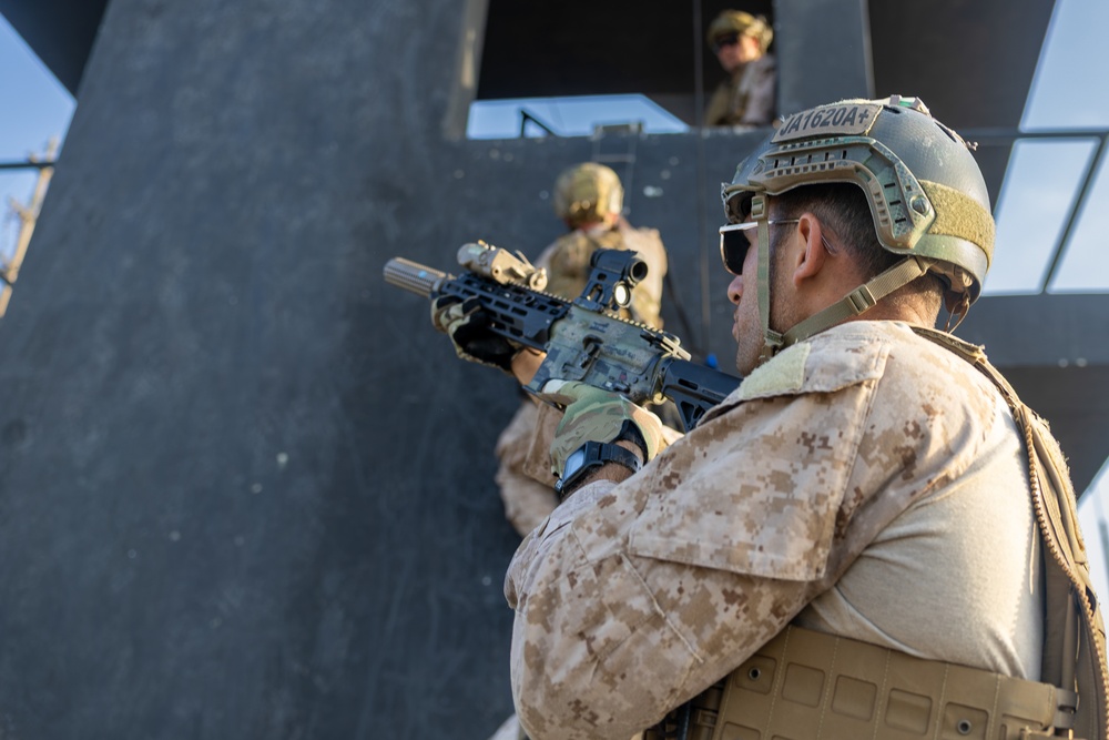 The image shows military personnel in desert camouflage uniforms conducting what appears to be a tactical training exercise or operation near a structure. The foreground soldier is armed with an assault rifle equipped with optics and is in a ready position. At least two additional personnel are visible ascending or positioned on a staircase structure in the background.