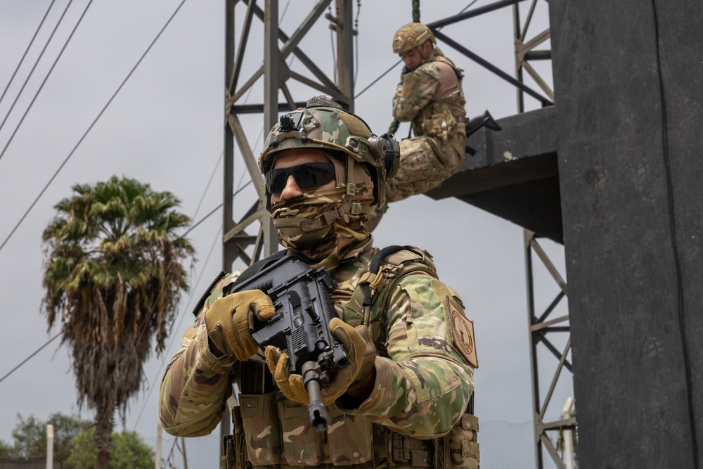 Two heavily armed and equipped military or special operations personnel are positioned near a metal tower structure. The foreground soldier is in full tactical gear including helmet, face covering, ballistic glasses, body armor, and is holding a compact assault rifle. A second soldier is visible in the background, positioned on an elevated platform of the metal structure.