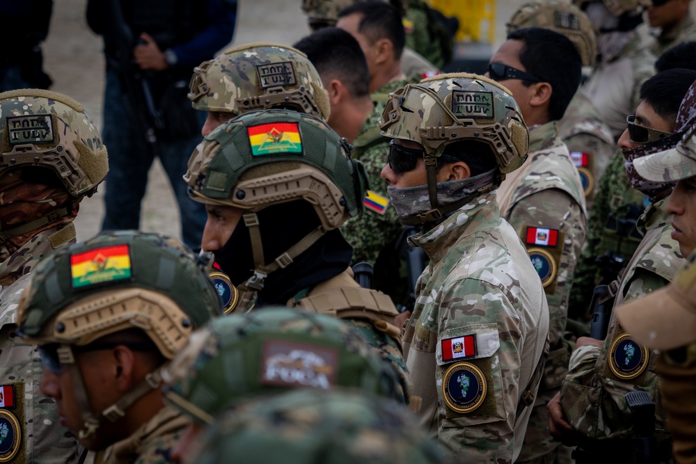 A group of military or special operations personnel wearing multicam/camouflage uniforms and tactical helmets are assembled together. Multiple national flag patches are visible on their uniforms and helmets, including Bolivia (red-yellow-green tricolor), Peru (red-white-red), and Ecuador (yellow-blue-red) flags, suggesting a multinational military exercise or joint operation. The personnel are equipped with tactical gear including ballistic helmets, face coverings, and sunglasses.
