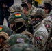 A group of military or special operations personnel wearing multicam/camouflage uniforms and tactical helmets are assembled together. Multiple national flag patches are visible on their uniforms and helmets, including Bolivia (red-yellow-green tricolor), Peru (red-white-red), and Ecuador (yellow-blue-red) flags, suggesting a multinational military exercise or joint operation. The personnel are equipped with tactical gear including ballistic helmets, face coverings, and sunglasses.