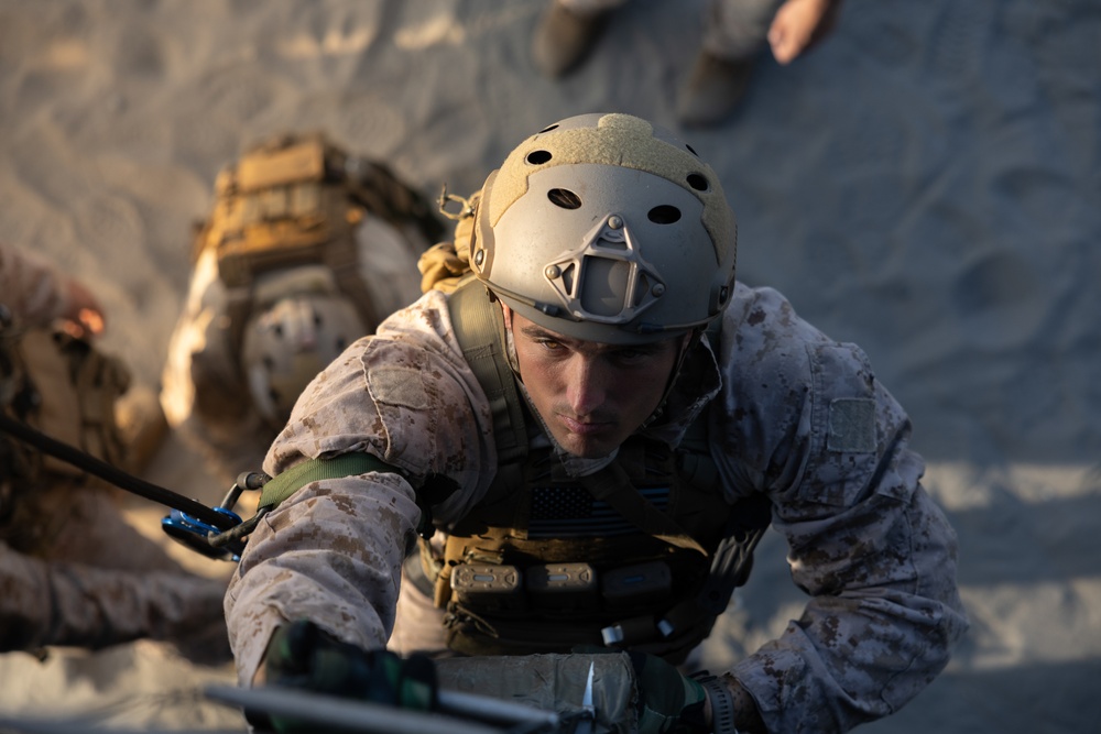 A military service member in desert MARPAT camouflage uniform is climbing or ascending what appears to be a sandy surface or structure using ropes and climbing equipment. The individual is wearing a tan FAST helmet with a visor mount and full tactical gear including a plate carrier vest. A second service member in similar gear is visible in the background, slightly out of focus.