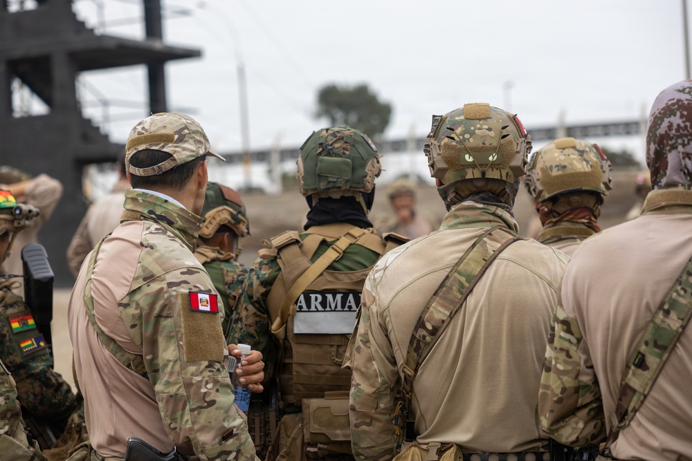 A group of military personnel in tactical gear are gathered together, appearing to be in a briefing or assembly. Multiple national flags are visible on uniforms, including what appears to be the Peruvian flag and Bolivian flag patches, suggesting a multinational military exercise or joint operation. The soldiers are equipped with helmets, body armor, and tactical vests.