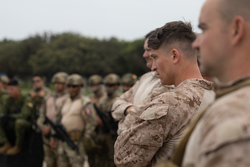 A group of military personnel from what appears to be multiple nations are assembled outdoors in a formation or briefing setting. The foreground shows soldiers in desert digital camouflage (consistent with USMC MARPAT desert pattern), while background personnel wear varied uniforms including woodland camouflage patterns. The setting appears to be a multinational military exercise or joint training event.