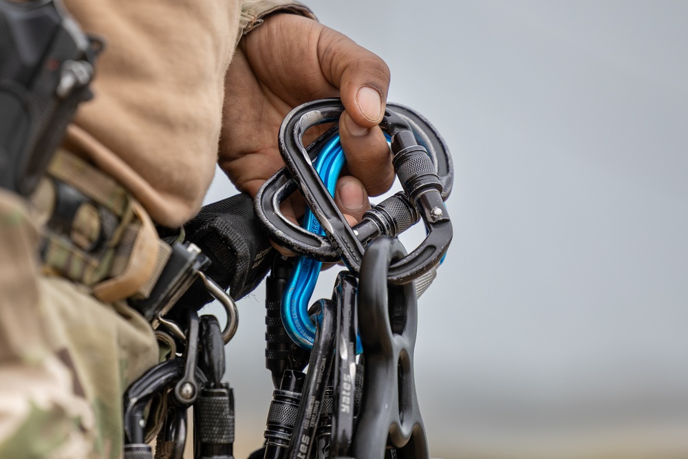 A person wearing military camouflage and a tan/beige sleeve is holding multiple carabiners in their hand. The carabiners are predominantly black/dark metal with one visibly blue carabiner among them. Additional carabiners and rigging equipment are visible attached to the person's gear on the left side of the frame.