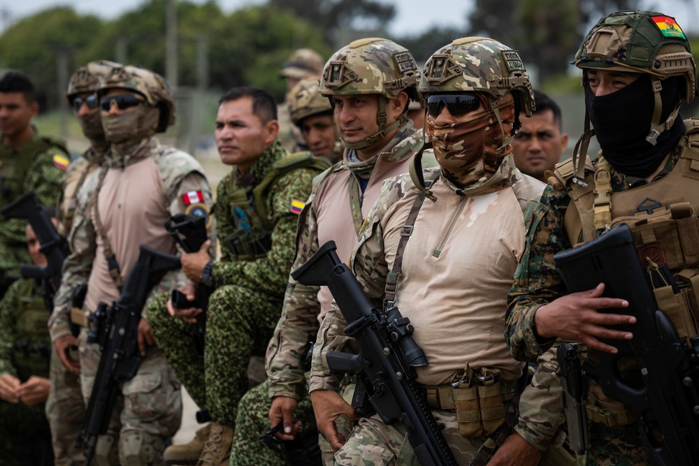 A group of military personnel from multiple nations are assembled together, likely during a joint military exercise or multinational training event. The soldiers display various national flags on their uniforms and wear a mix of tactical gear, helmets, and face coverings. The setting appears to be an outdoor military installation or training ground.