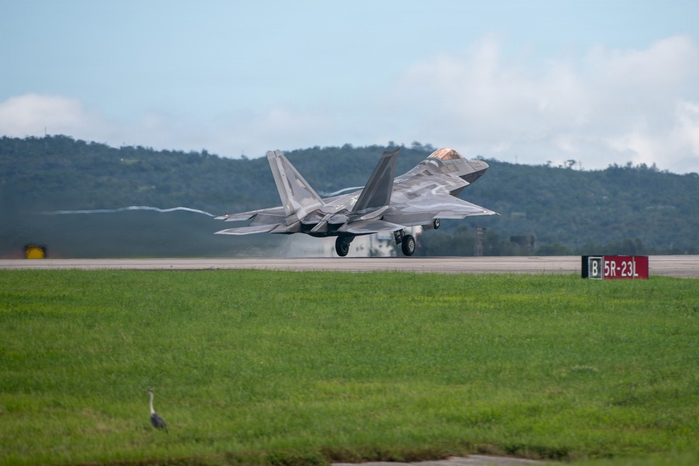 An F-22 Raptor stealth fighter jet is captured during takeoff or landing on a military airstrip, with landing gear still deployed and visible vapor trails streaming from the wingtips. The aircraft is painted in a gray camouflage scheme and the runway signage reads '5R-23L', indicating a specific runway designation. A heron bird is visible on the grass infield in the foreground.