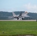 An F-22 Raptor stealth fighter jet is captured during takeoff or landing on a military airstrip, with landing gear still deployed and visible vapor trails streaming from the wingtips. The aircraft is painted in a gray camouflage scheme and the runway signage reads '5R-23L', indicating a specific runway designation. A heron bird is visible on the grass infield in the foreground.