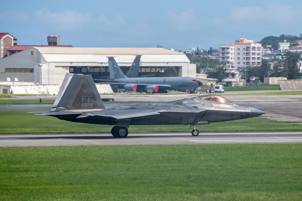 A U.S. Air Force F-22 Raptor stealth fighter jet with tail code 'FF' and serial number 078 is taxiing on a runway. In the background, a large tanker aircraft (likely a KC-135) with tail number 22 is parked near a hangar. The setting appears to be a military airbase surrounded by urban infrastructure and vegetation, consistent with a base in the Pacific region.