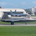 A U.S. Air Force F-22 Raptor stealth fighter jet with tail code 'FF' and serial number 078 is taxiing on a runway. In the background, a large tanker aircraft (likely a KC-135) with tail number 22 is parked near a hangar. The setting appears to be a military airbase surrounded by urban infrastructure and vegetation, consistent with a base in the Pacific region.