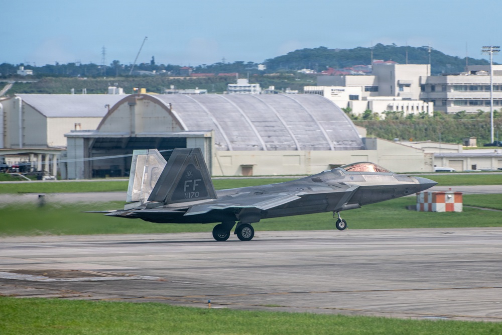 An F-22 Raptor stealth fighter jet is taxiing on a runway at a military air base. The aircraft displays tail code 'FF' and serial number '170', indicating it belongs to the 1st Fighter Wing based at Langley Air Force Base. The background features large aircraft hangars, urban infrastructure, and tree-covered hills, suggesting a location in a densely populated area, consistent with Kadena Air Base in Okinawa, Japan.