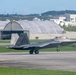 An F-22 Raptor stealth fighter jet is taxiing on a runway at a military air base. The aircraft displays tail code 'FF' and serial number '170', indicating it belongs to the 1st Fighter Wing based at Langley Air Force Base. The background features large aircraft hangars, urban infrastructure, and tree-covered hills, suggesting a location in a densely populated area, consistent with Kadena Air Base in Okinawa, Japan.