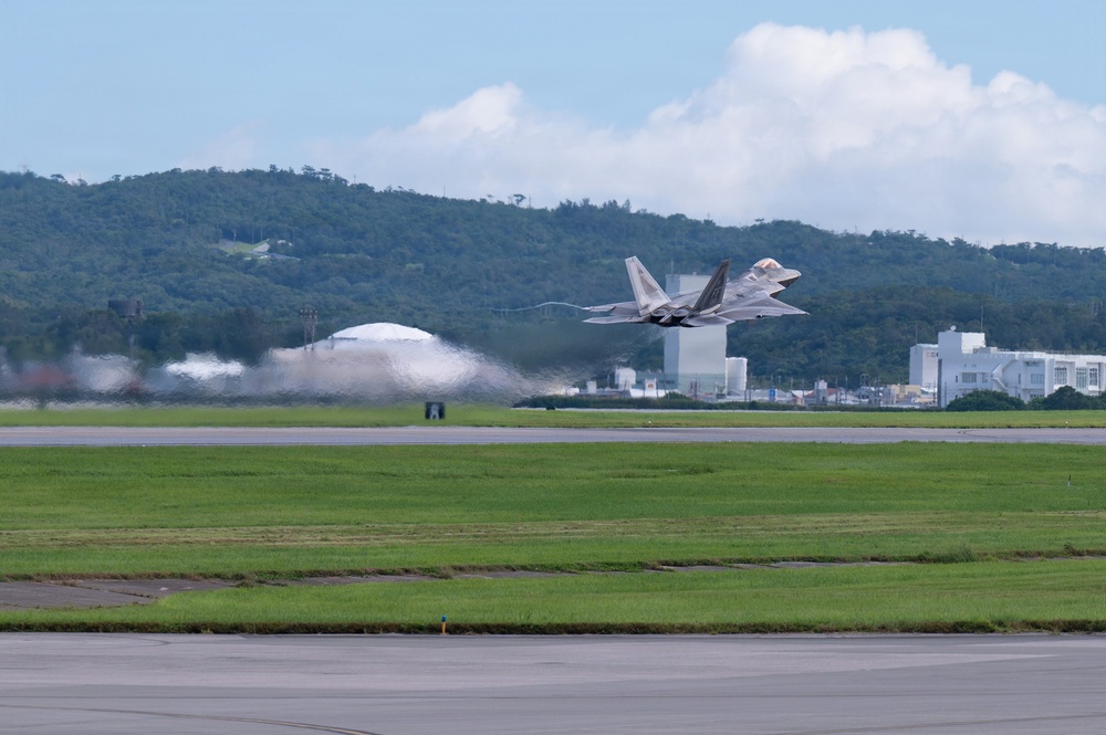 A military fighter jet, consistent with an F-22 Raptor, is captured in the moment of takeoff from a runway at what appears to be a military airbase. The aircraft is banking slightly as it lifts off, with heat distortion and jet wash visible behind it. The base is surrounded by green, forested hills and features several white buildings and a dome-shaped hangar structure in the background.