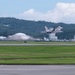 A military fighter jet, consistent with an F-22 Raptor, is captured in the moment of takeoff from a runway at what appears to be a military airbase. The aircraft is banking slightly as it lifts off, with heat distortion and jet wash visible behind it. The base is surrounded by green, forested hills and features several white buildings and a dome-shaped hangar structure in the background.