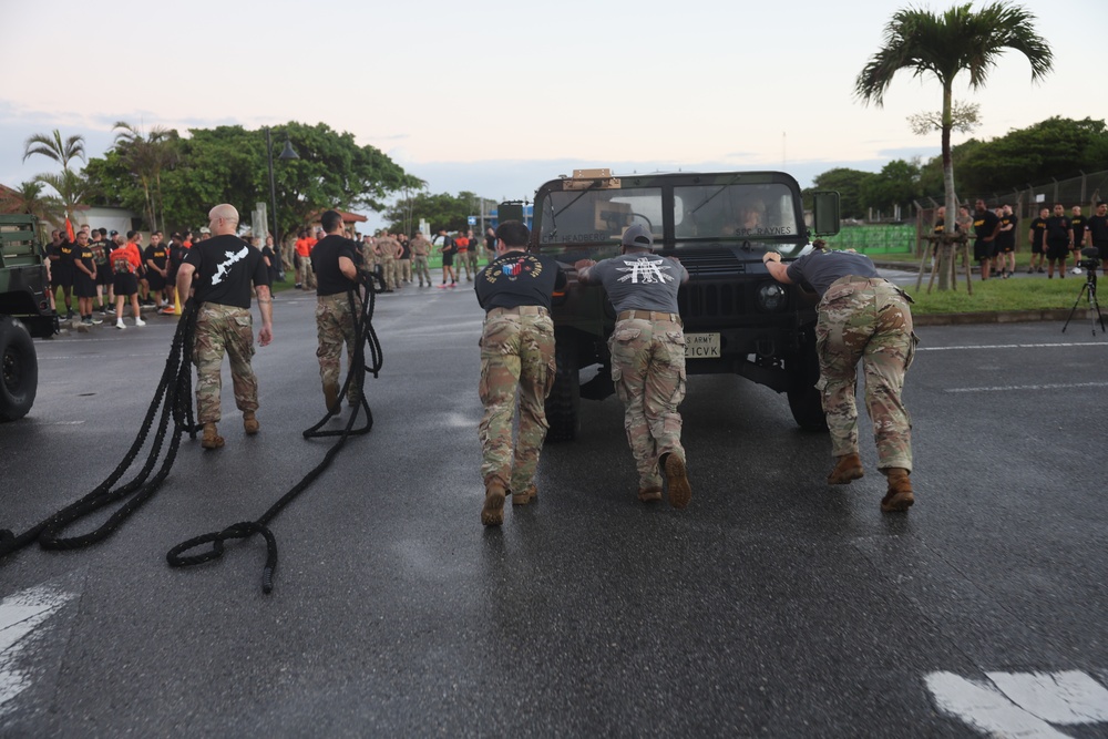 Military personnel in camouflage uniforms and black t-shirts are pushing and pulling a U.S. Army Humvee on a wet road surface, likely during a physical fitness or team-building exercise. The vehicle bears a U.S. Army license plate partially visible as 'Z1CVK'. A crowd of uniformed observers lines the sides of the road, watching the activity in what appears to be a tropical military installation.