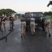 Military personnel in camouflage uniforms and black t-shirts are pushing and pulling a U.S. Army Humvee on a wet road surface, likely during a physical fitness or team-building exercise. The vehicle bears a U.S. Army license plate partially visible as 'Z1CVK'. A crowd of uniformed observers lines the sides of the road, watching the activity in what appears to be a tropical military installation.