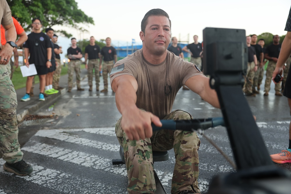 A military service member in MultiCam/OCP camouflage uniform is using a rowing machine (ergometer) on an outdoor paved surface, likely during a fitness assessment or competition. A crowd of uniformed personnel and some civilians stand in the background observing the activity. The setting appears to be a military installation, possibly in a tropical or subtropical location based on the vegetation.