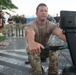 A military service member in MultiCam/OCP camouflage uniform is using a rowing machine (ergometer) on an outdoor paved surface, likely during a fitness assessment or competition. A crowd of uniformed personnel and some civilians stand in the background observing the activity. The setting appears to be a military installation, possibly in a tropical or subtropical location based on the vegetation.