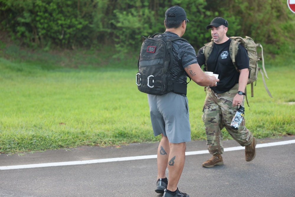 Two men are walking on a paved road, both carrying backpacks. The man in the foreground wears civilian athletic clothing with a tactical-style backpack, while the man in the background wears military camouflage pants, a black t-shirt, and carries a military-style rucksack. They appear to be participating in a ruck march or similar fitness/military event.