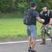 Two men are walking on a paved road, both carrying backpacks. The man in the foreground wears civilian athletic clothing with a tactical-style backpack, while the man in the background wears military camouflage pants, a black t-shirt, and carries a military-style rucksack. They appear to be participating in a ruck march or similar fitness/military event.