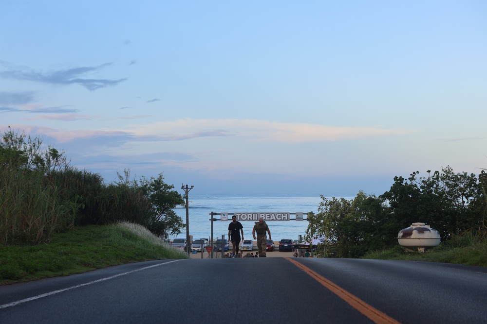 A road leads toward a beach entrance marked with a sign reading 'TORII BEACH,' with the ocean visible in the background under a twilight sky. Two individuals are walking toward the camera on the road, and a small parking area with vehicles is visible near the entrance gate. The scene appears to be at a military recreational beach facility, likely on Okinawa, Japan.
