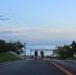 A road leads toward a beach entrance marked with a sign reading 'TORII BEACH,' with the ocean visible in the background under a twilight sky. Two individuals are walking toward the camera on the road, and a small parking area with vehicles is visible near the entrance gate. The scene appears to be at a military recreational beach facility, likely on Okinawa, Japan.