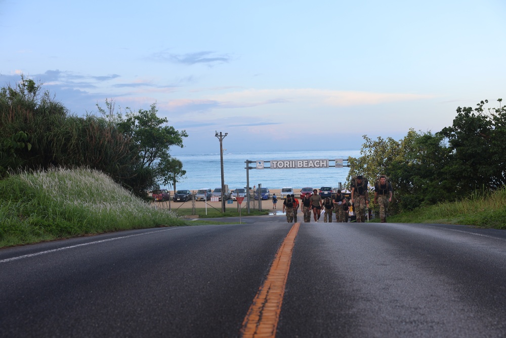 A road leads toward Torii Beach, a military recreational facility, with a group of individuals in camouflage uniforms walking away from the beach toward the camera. The scene is captured at dusk or dawn, with a calm ocean visible in the background and lush tropical vegetation on both sides of the road. Several vehicles are parked near the beach entrance.