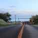 A road leads toward Torii Beach, a military recreational facility, with a group of individuals in camouflage uniforms walking away from the beach toward the camera. The scene is captured at dusk or dawn, with a calm ocean visible in the background and lush tropical vegetation on both sides of the road. Several vehicles are parked near the beach entrance.