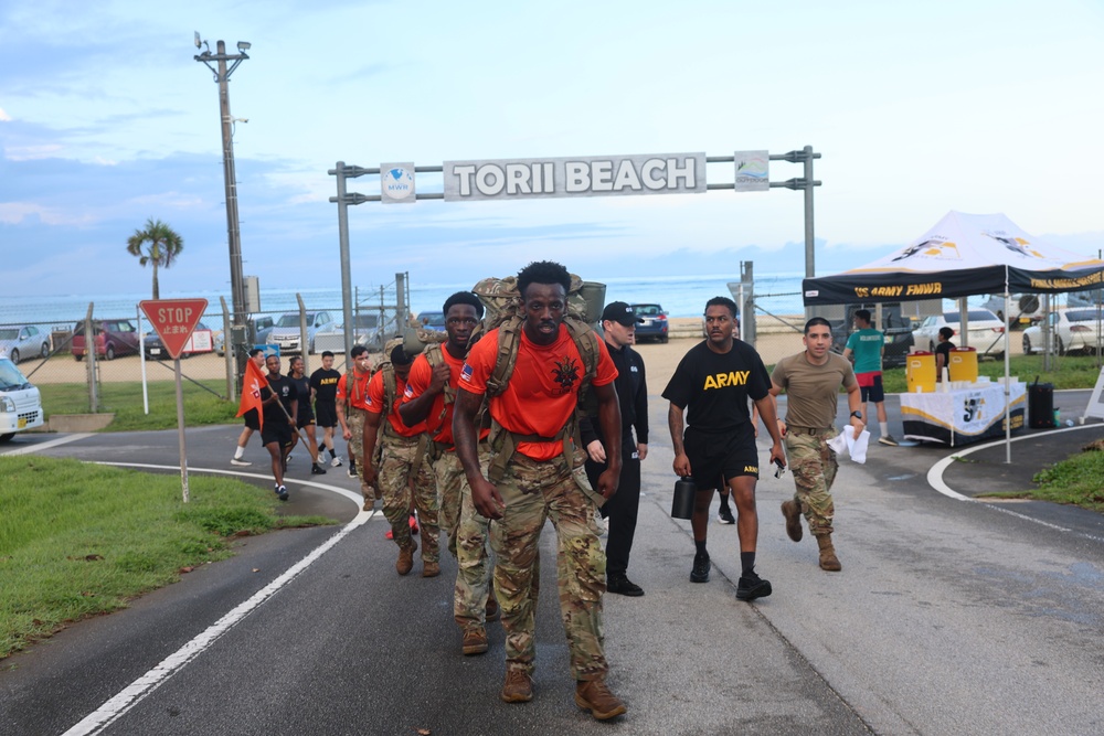 A group of U.S. Army soldiers are marching away from Torii Beach, located on what appears to be Torii Station in Okinawa, Japan. The lead soldiers are wearing orange shirts with camouflage pants and carrying rucksacks, suggesting a ruck march or physical fitness event. A U.S. Army MWR (Morale, Welfare, and Recreation) tent is visible to the right, indicating an organized military event.