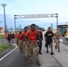 A group of U.S. Army soldiers are marching away from Torii Beach, located on what appears to be Torii Station in Okinawa, Japan. The lead soldiers are wearing orange shirts with camouflage pants and carrying rucksacks, suggesting a ruck march or physical fitness event. A U.S. Army MWR (Morale, Welfare, and Recreation) tent is visible to the right, indicating an organized military event.