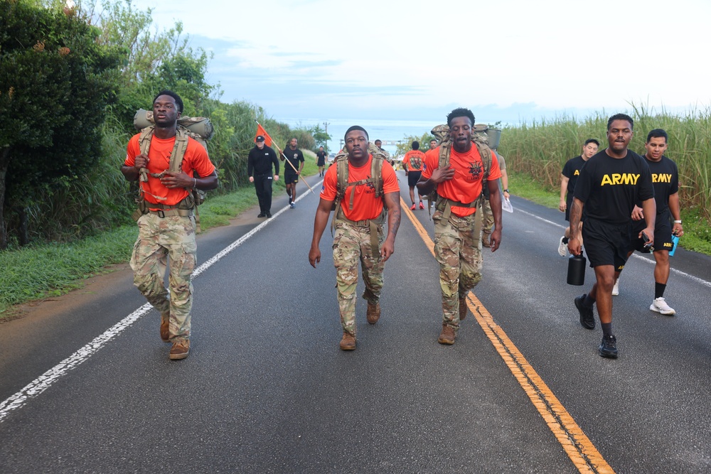 A group of U.S. Army soldiers and personnel are conducting a ruck march or physical training event along a paved road in what appears to be a tropical or coastal location. Three soldiers in the foreground wear orange shirts with camouflage pants and carry heavy rucksacks, while others in black Army PT uniforms accompany them. The setting features lush vegetation and a body of water visible in the background.