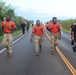A group of U.S. Army soldiers and personnel are conducting a ruck march or physical training event along a paved road in what appears to be a tropical or coastal location. Three soldiers in the foreground wear orange shirts with camouflage pants and carry heavy rucksacks, while others in black Army PT uniforms accompany them. The setting features lush vegetation and a body of water visible in the background.