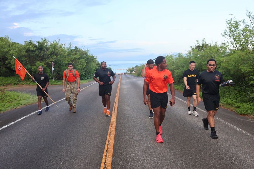 A group of U.S. Army soldiers are running and walking along a two-lane road lined with green trees, with a body of water visible in the background. Most are wearing black Army PT (Physical Training) shirts and shorts, while one soldier in the center wears an orange shirt and another is in full camouflage uniform carrying a unit guidon flag. This appears to be a military physical training run or ruck march event.