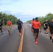 A group of U.S. Army soldiers are running and walking along a two-lane road lined with green trees, with a body of water visible in the background. Most are wearing black Army PT (Physical Training) shirts and shorts, while one soldier in the center wears an orange shirt and another is in full camouflage uniform carrying a unit guidon flag. This appears to be a military physical training run or ruck march event.