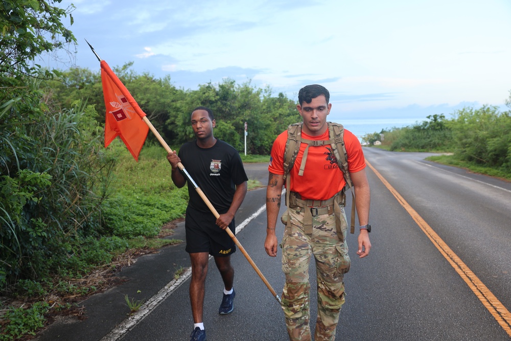 Two U.S. Army soldiers are marching along a paved road in what appears to be a tropical or subtropical environment. The soldier in the foreground wears an orange shirt, camouflage pants, and carries a tactical backpack, while the soldier behind him carries an orange military guidon flag. The setting suggests a ruck march or unit physical training event.
