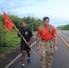 Two U.S. Army soldiers are marching along a paved road in what appears to be a tropical or subtropical environment. The soldier in the foreground wears an orange shirt, camouflage pants, and carries a tactical backpack, while the soldier behind him carries an orange military guidon flag. The setting suggests a ruck march or unit physical training event.