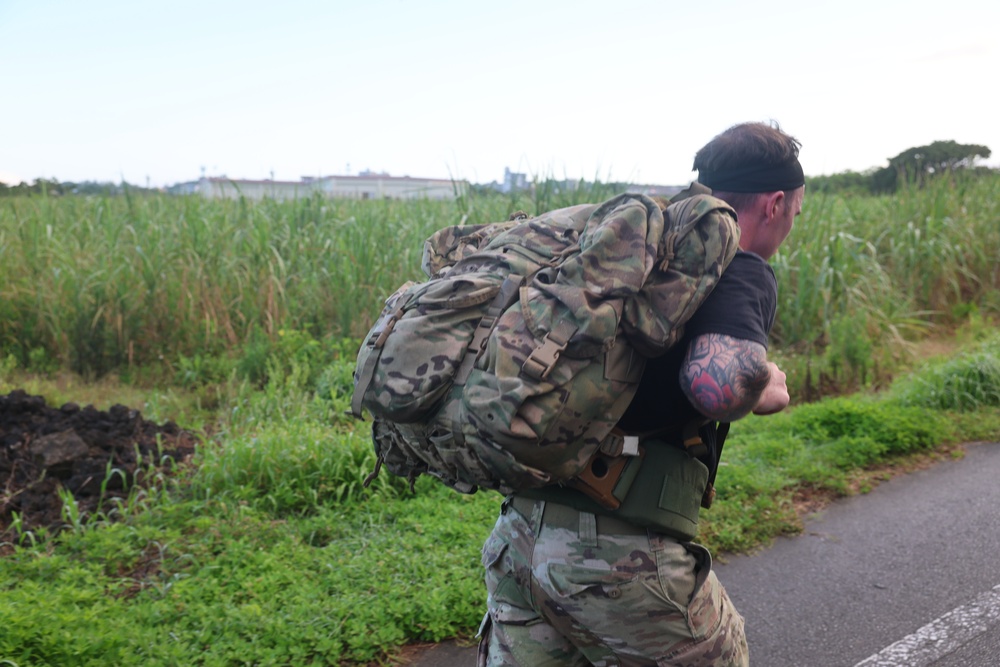 A male individual wearing military-style camouflage clothing is carrying a large, heavily loaded tactical rucksack while moving along a paved road. He is wearing a black t-shirt, camouflage pants, a green belt, and a black headband, with a visible tattoo on his right arm. The background features dense tall grass, green vegetation, and distant buildings under an overcast sky.