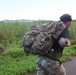 A male individual wearing military-style camouflage clothing is carrying a large, heavily loaded tactical rucksack while moving along a paved road. He is wearing a black t-shirt, camouflage pants, a green belt, and a black headband, with a visible tattoo on his right arm. The background features dense tall grass, green vegetation, and distant buildings under an overcast sky.