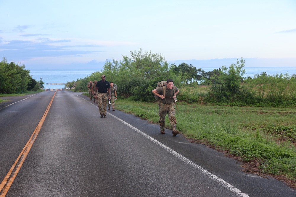 A group of military personnel in camouflage uniforms are conducting a ruck march along a two-lane coastal road. One soldier in the foreground is running with a heavy backpack while others walk behind, led by a person in a black shirt. The setting appears to be a tropical or subtropical coastal area, likely during early morning based on the soft lighting.