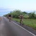 A group of military personnel in camouflage uniforms are conducting a ruck march along a two-lane coastal road. One soldier in the foreground is running with a heavy backpack while others walk behind, led by a person in a black shirt. The setting appears to be a tropical or subtropical coastal area, likely during early morning based on the soft lighting.