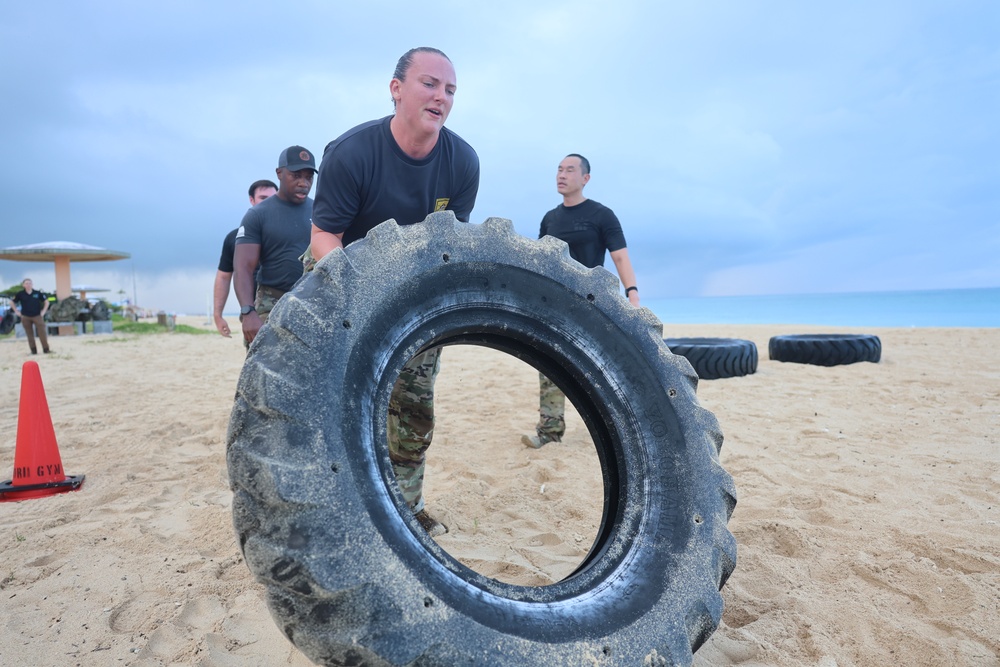 A group of individuals in black military-style uniforms are conducting a physical fitness training exercise on a sandy beach. The primary subject, a woman in the foreground, is flipping or pushing a large, sand-covered tractor tire. Additional participants and large tires are visible in the background near the ocean.