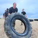 A group of individuals in black military-style uniforms are conducting a physical fitness training exercise on a sandy beach. The primary subject, a woman in the foreground, is flipping or pushing a large, sand-covered tractor tire. Additional participants and large tires are visible in the background near the ocean.