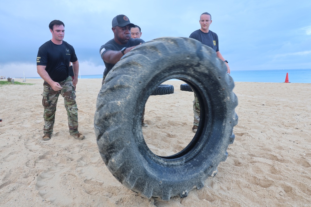 A group of military personnel are conducting a physical training exercise on a sandy beach. One individual is flipping a large, heavy tractor tire while others observe or assist. The setting appears to be an early morning or overcast day near an ocean shoreline.