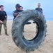 A group of military personnel are conducting a physical training exercise on a sandy beach. One individual is flipping a large, heavy tractor tire while others observe or assist. The setting appears to be an early morning or overcast day near an ocean shoreline.