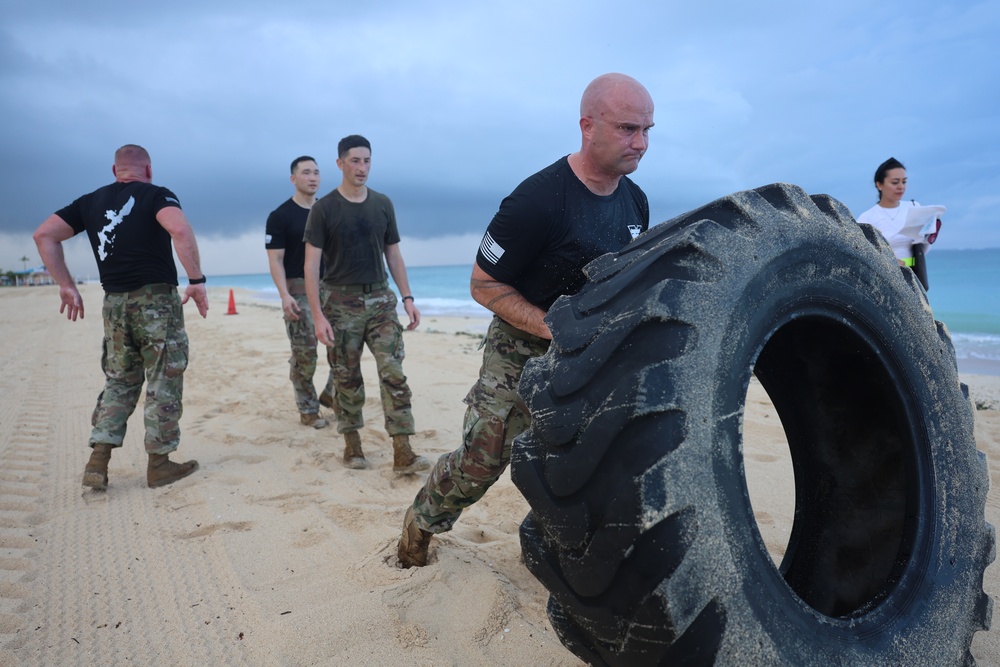 A group of individuals in military-style uniforms are conducting a physical fitness training exercise on a sandy beach. The central figure, a bald man in a black t-shirt and camouflage pants, is performing a large tire flip. Several other participants and an observer with a clipboard are visible in the background near the shoreline.