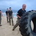 A group of individuals in military-style uniforms are conducting a physical fitness training exercise on a sandy beach. The central figure, a bald man in a black t-shirt and camouflage pants, is performing a large tire flip. Several other participants and an observer with a clipboard are visible in the background near the shoreline.