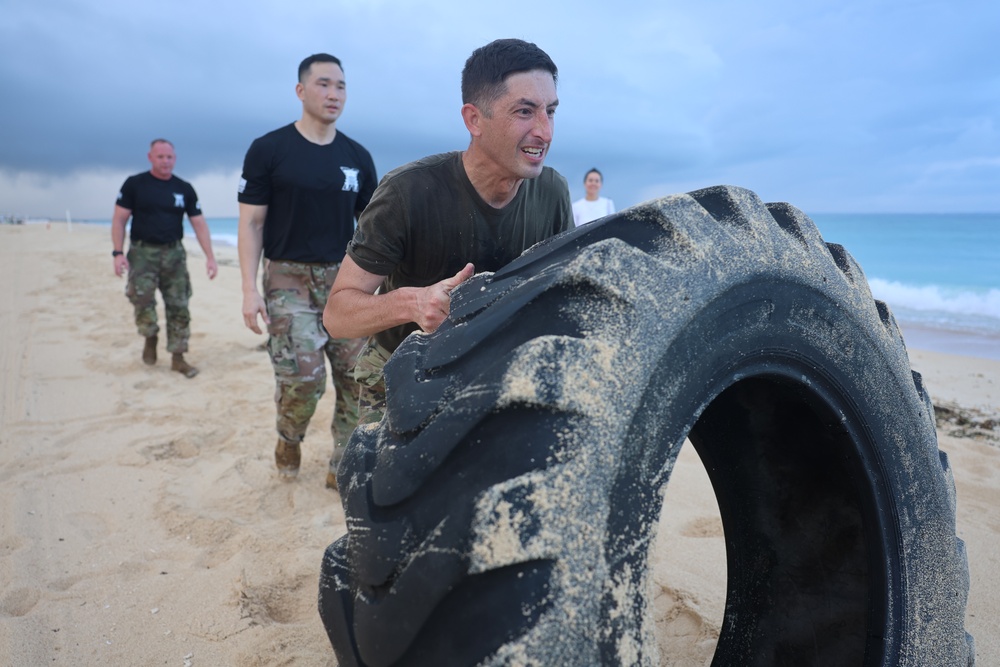 A man in military-style clothing is flipping a large, sand-covered tractor tire on a beach during what appears to be a physical fitness training exercise. Several other individuals in military and civilian attire are visible in the background observing or participating. The setting is an overcast beach with ocean waves visible in the background.