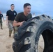 A man in military-style clothing is flipping a large, sand-covered tractor tire on a beach during what appears to be a physical fitness training exercise. Several other individuals in military and civilian attire are visible in the background observing or participating. The setting is an overcast beach with ocean waves visible in the background.